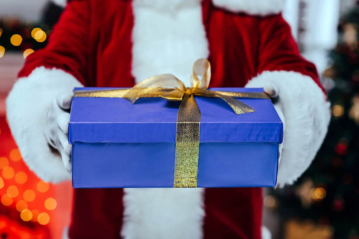 Holiday Gift A person in a red Santa suit holds a blue gift box with a gold ribbon in front of a festive background with blurred lights.