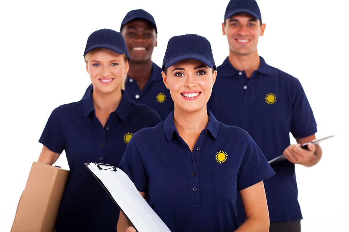 Staff Uniforms A group of four smiling individuals in new New Braunfels custom apparel in navy blue polo shirts and caps, two holding clipboards and one carrying a box, standing together against a white background.